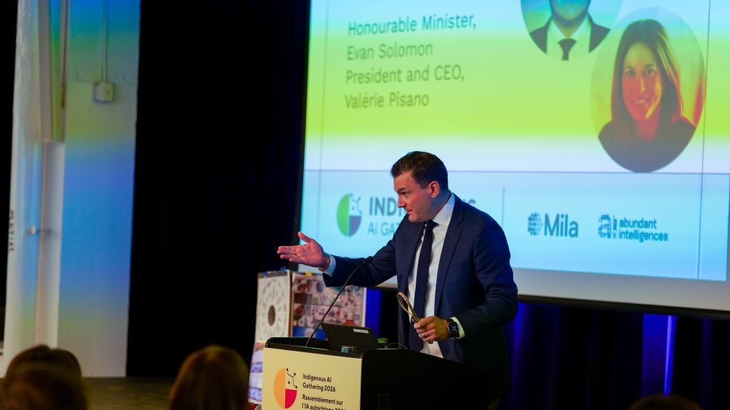 Evan Solomon, ministro canadense de Inteligência Artificial e Inovação Digital, discursa em um púlpito durante o Encontro Indígena de IA 2026. Ao fundo, são projetadas as logomarcas do evento, do Mila e da Abundant Intelligences.